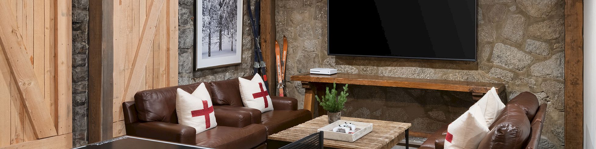 A cozy stone-walled game room with a ping-pong table, brown leather sofas, framed photos, a fireplace, and a large wall TV.