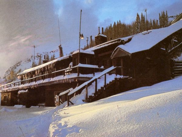 A snowy freight train crosses a bridge through a forest of evergreens, with snow-covered rails and buildings in view.