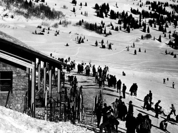 Black-and-white ski scene: crowds of skiers line up on a lift platform beside a stone building overlooking snowy slopes.