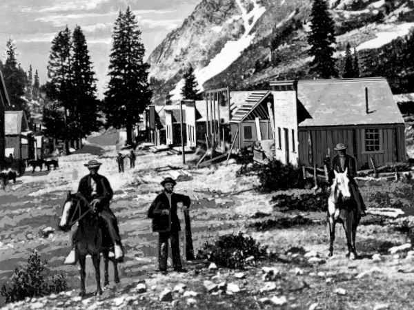 In a snowy Alpine landscape, two men stand outside damaged barracks/cabins with mountains and tall pine trees in the background.