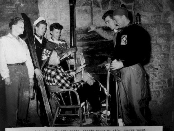 A black-and-white scene of several people in a room as a man in uniform leans in to talk beside a seated chair and equipment.