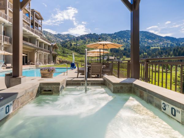 An outdoor heated pool and hot tub area with lounge chairs and umbrellas, overlooking green mountain views and apartment balconies.