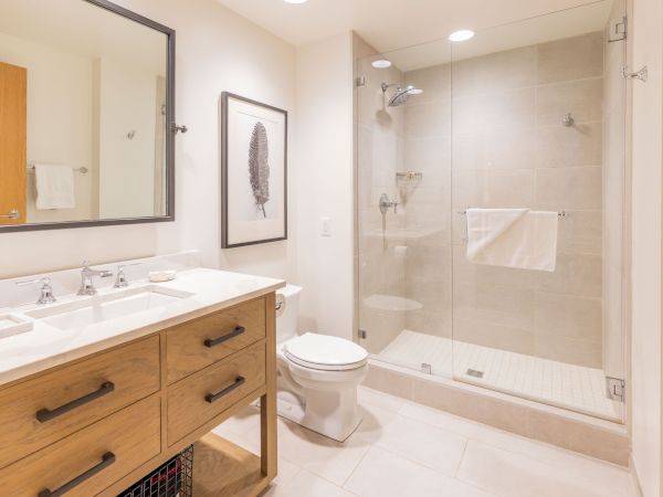 A bright modern bathroom with a double-sink vanity, framed mirror, toilet, and glass-enclosed walk-in shower.