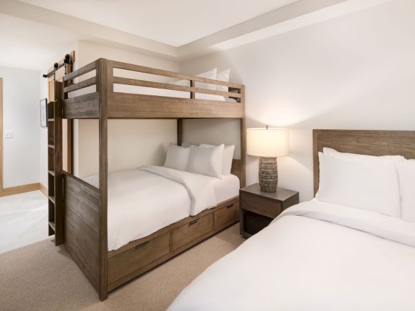 A modern bedroom with a wooden bunk bed, two beds with white linens, a bedside lamp, and beige walls.