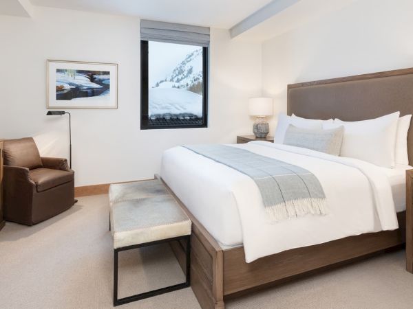 A cozy modern bedroom with a large bed, nightstands, pillows, and a chair, plus a snowy mountain view through the window.