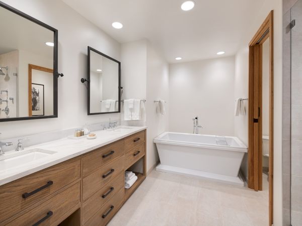 Bright modern bathroom with double sinks, wood vanity, large mirrors, towels, and a white soaking bathtub.
