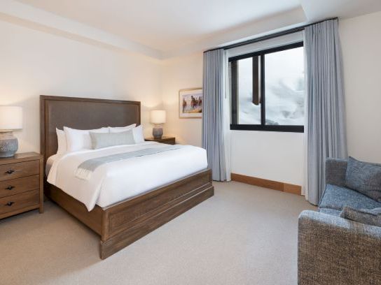 A modern hotel bedroom with a large bed, bedside tables and lamps, and a window with curtains beside a small sofa.