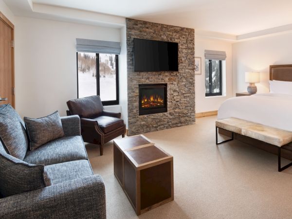 A cozy room with a stone fireplace, sofa and chair, coffee tables, a bed, and snowy mountain views through large windows.