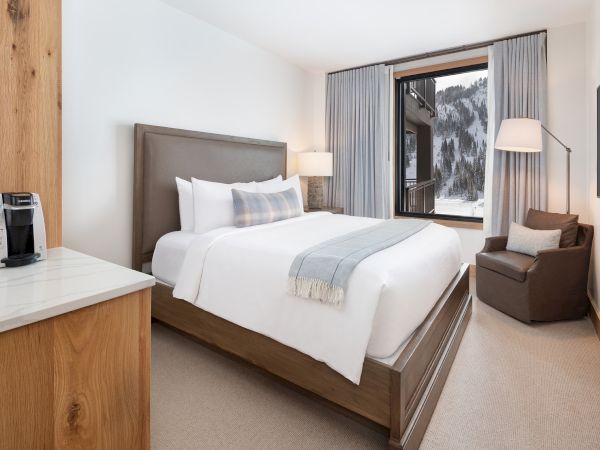 A modern hotel bedroom with a neatly made bed, curtains, mountain view, lamps, lounge chair, and a Keurig on the counter.
