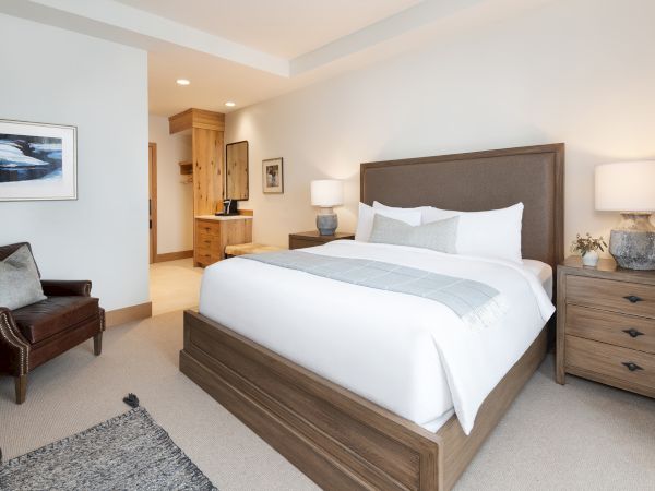 A bright, modern bedroom with a large bed, gray headboard, bedside lamps, wooden dresser, and a cozy seating chair by the window.
