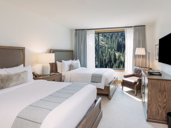 A modern hotel room with two beds, bedside lamps, an armchair, a wall TV, and curtains framing a forested mountain view.
