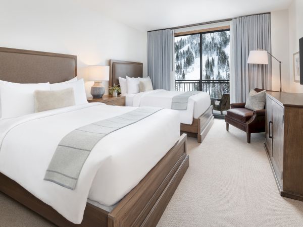 A bright hotel bedroom with two beds, gray curtains, a snowy forest view, a lamp, and a TV.