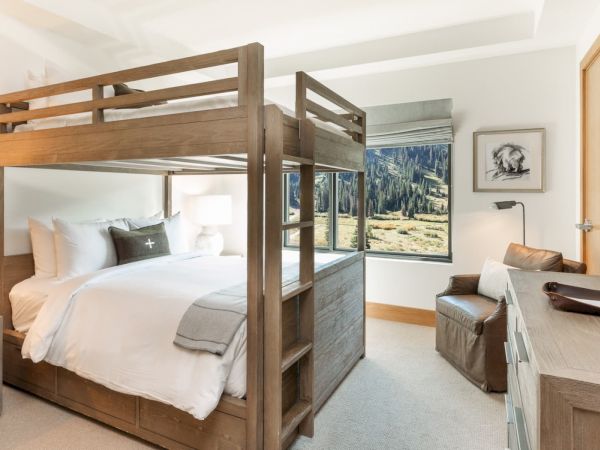 A modern hotel room with a wooden bunk bed, sofa, desk, and mountain view through large windows.