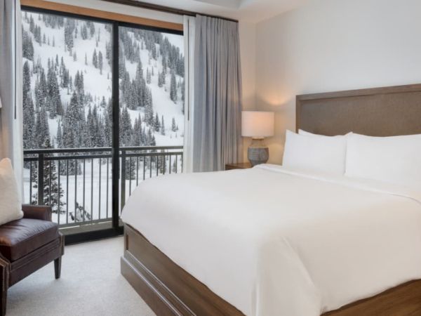 A cozy hotel bedroom with a large bed, lamps, chairs, and curtains, overlooking a snowy pine forest through tall windows.