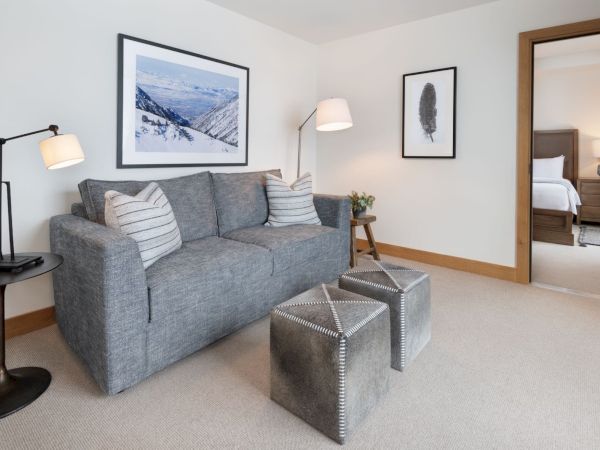 A bright living room with a gray sofa, pillows and ottomans, floor lamp, framed art, and a glimpse of a bedroom.