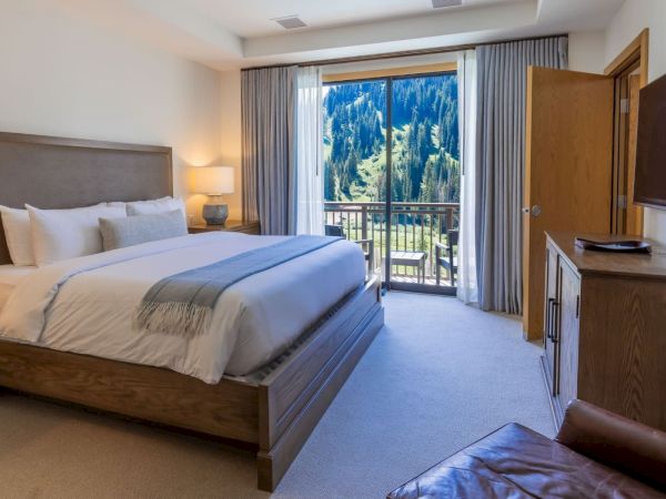 A modern hotel bedroom with a large bed, TV and cabinets, and a balcony view of tall blue pine-covered mountains.