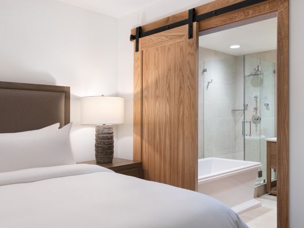 A modern bedroom with a wooden door open to a clean bathroom, showing a glass shower and white bathtub.