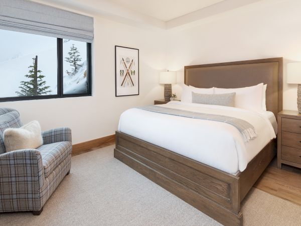 A modern bedroom with a wooden bed, lamps, plaid armchair, framed art, and a large window showing snowy mountains and pine trees.