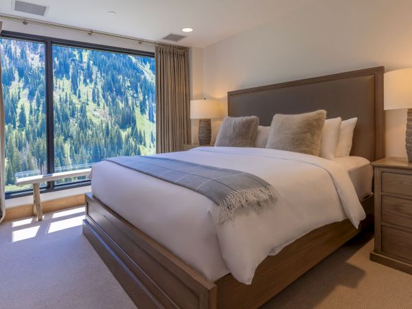 A cozy hotel bedroom with a neatly made bed, soft curtains, and a large window framing pine-covered mountains in view.