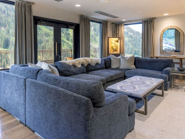 A modern living room with a blue sectional sofa, patterned ottoman, tall curtains, and mountain views through large windows.
