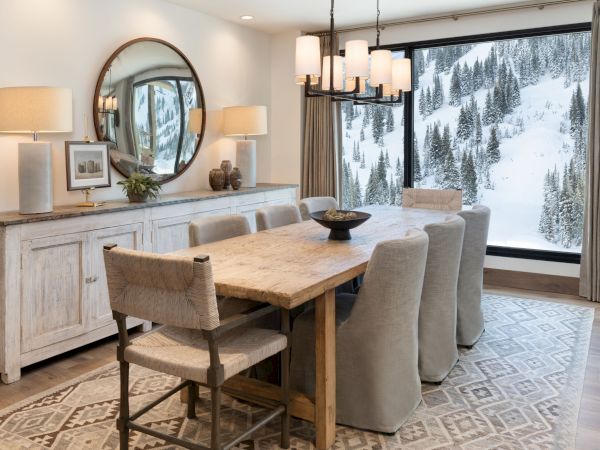 A cozy alpine dining room with a rustic wood table, upholstered chairs, pendant lights, and snowy forest views through large windows.