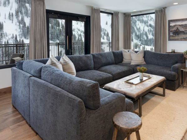 A cozy living room with a blue sectional, coffee table, curtains, and large windows showing snowy pine forests outside.