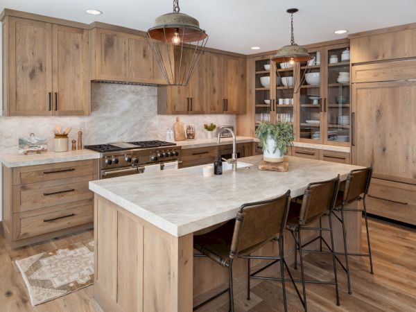 A rustic kitchen with wood cabinets, a marble island, stove and sink, pendant lights, and a bar with stools.