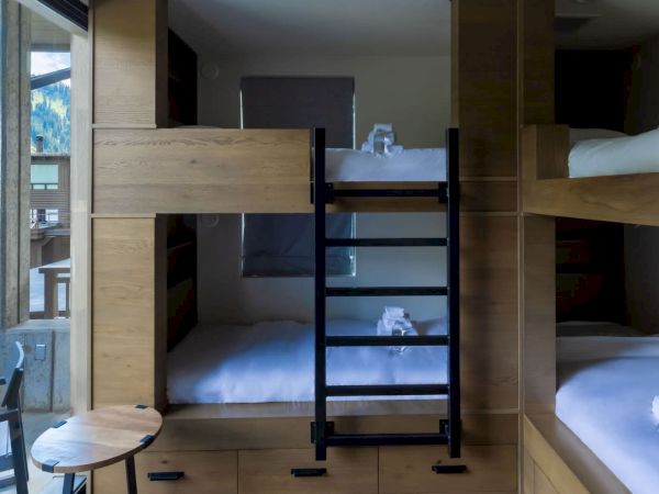 A modern alpine hotel room with built-in bunk beds, wooden panels, white linens, a small round table, and mountain-view windows.