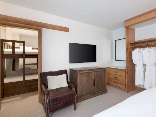 A modern hotel suite shows a TV on a wooden dresser, a lounge chair, and two hanging white bathrobes.