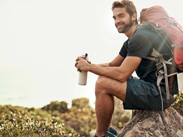 A smiling hiker with a red backpack drinks from a bottle while resting on a rock outdoors, surrounded by nature.