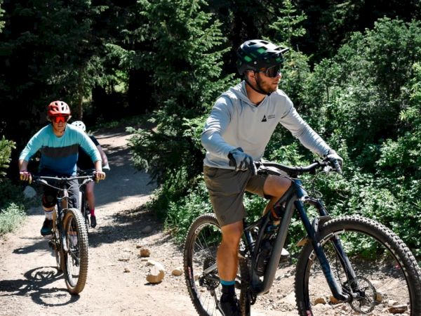 Two mountain bikers ride along a rocky forest trail, wearing helmets and riding hardtails under sunny greenery.