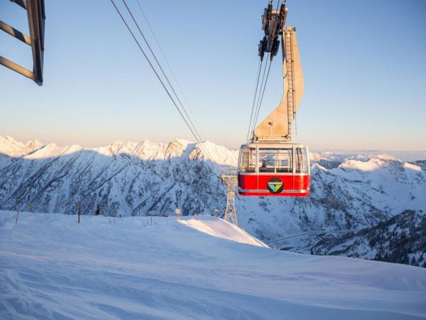A red-and-white ski gondola hangs on cables above snowy ski slopes with mountains in the background, at dusk.