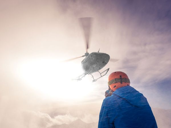 A medic in winter gear stands in the clouds as a military helicopter hovers overhead, ready to rescue.