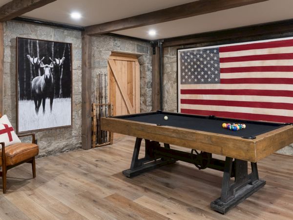 A rustic game room with a billiards table, a U.S. flag wall banner, and deer artwork on stone walls, plus a leather chair.
