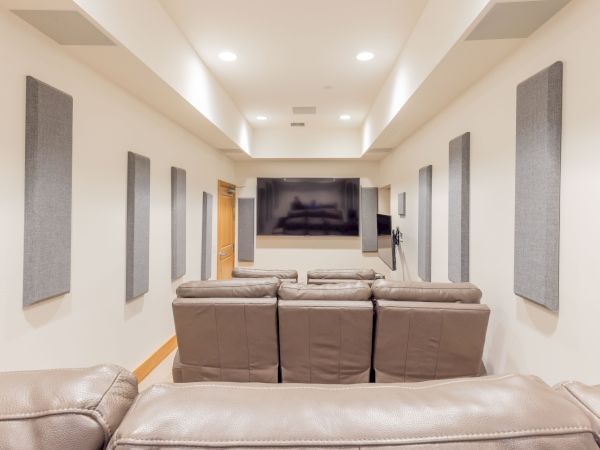 A home theater room with a gray recliner sofa, wall-mounted TV, acoustic panels, and recessed ceiling lights.