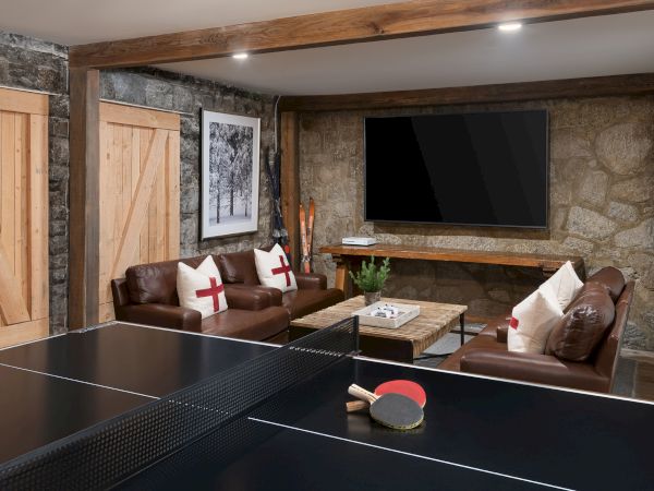 A modern lounge with a ping-pong table, leather sofas, stone fireplace wall, framed photos, and a large wall TV.