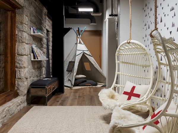 A cozy hallway with stone walls, a large rug, a wooden chest, a white wicker hanging chair with red cross cushions, and a teepee bed.