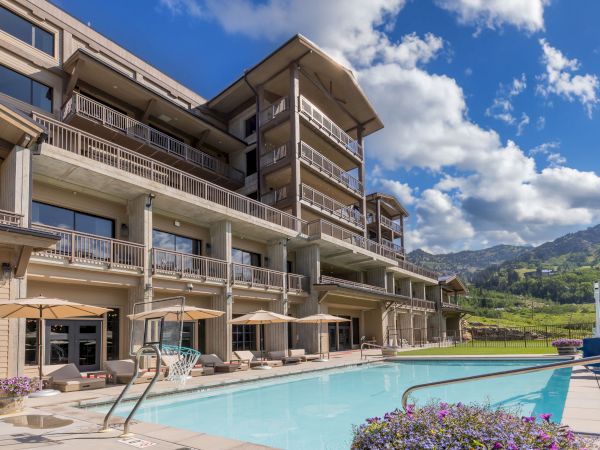 Balcony-lined hotel building beside a sparkling outdoor swimming pool, with umbrellas and green hills under a bright blue sky.
