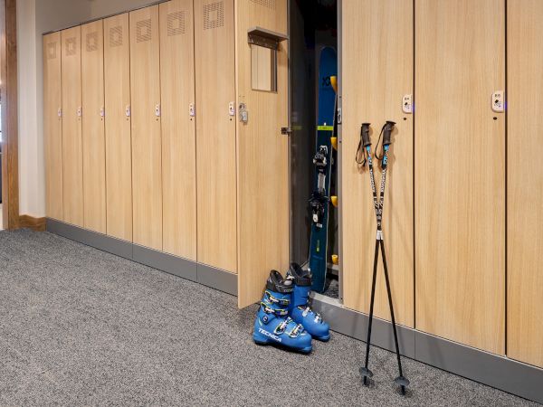 Ski boots and poles sit in open indoor storage lockers with numbered doors and call buttons along a hallway.