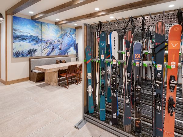 Indoor ski rack room with many colorful skis on metal pegs, plus a bench and mountain mural on the wall.