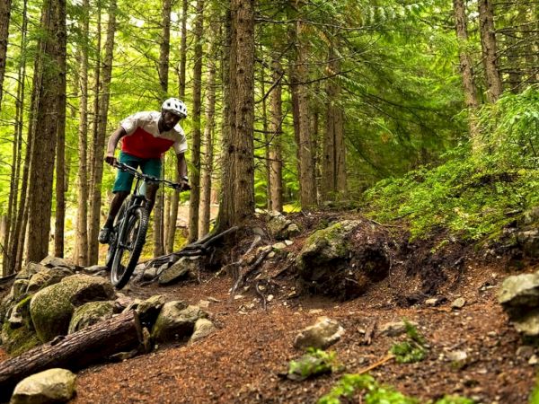 A mountain biker rides a rocky downhill trail through a green forest, navigating between tall trees.