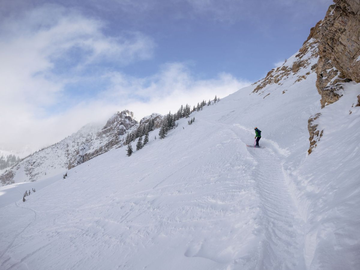 A skier in bright gear carves down a snowy alpine slope beside rocky cliffs under a blue sky.