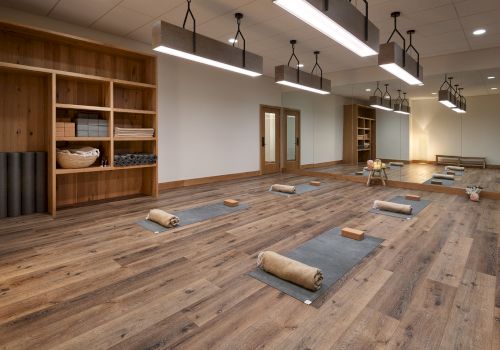 A bright yoga studio with wooden storage shelves, floor mats, folded towels, yoga blocks, and hanging pendant lights.