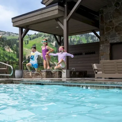 A woman and three kids play at an outdoor pool beside a mountain lodge, splashing in the clear water.