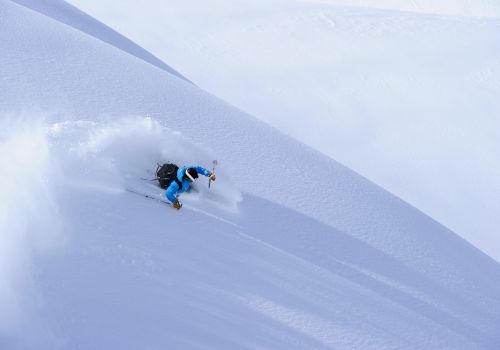 A skier in blue jackets carves through deep powder on a steep mountain slope, kicking up snow with poles.
