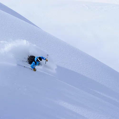 A skier in blue jackets carves through deep powder on a steep mountain slope, kicking up snow with poles.