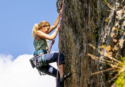 A climber in harness and chalk bag grips a rocky cliff face, climbing with colorful ropes under a clear blue sky.