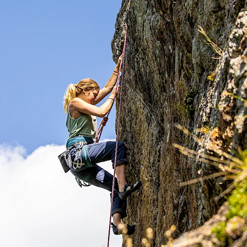 A climber in harness and chalk bag grips a rocky cliff face, climbing with colorful ropes under a clear blue sky.