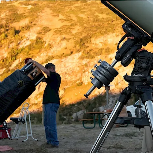 A group sets up and adjusts a large telescope outdoors on a hillside at sunset.
