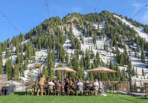 A sunny ski resort scene with chairlift cables over snowy mountains, tall pines, and people dining outdoors under umbrellas.